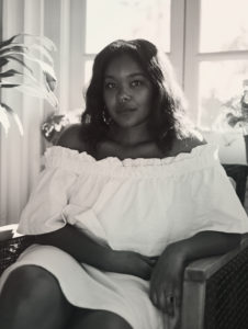 This is a Black and white headshot of Courtney Faye Taylor. She sis seated beside a house plant and in front of lit window. She is wearing a white dress and hoop earrings. Shis is looking directly at the camera and has dark hair.